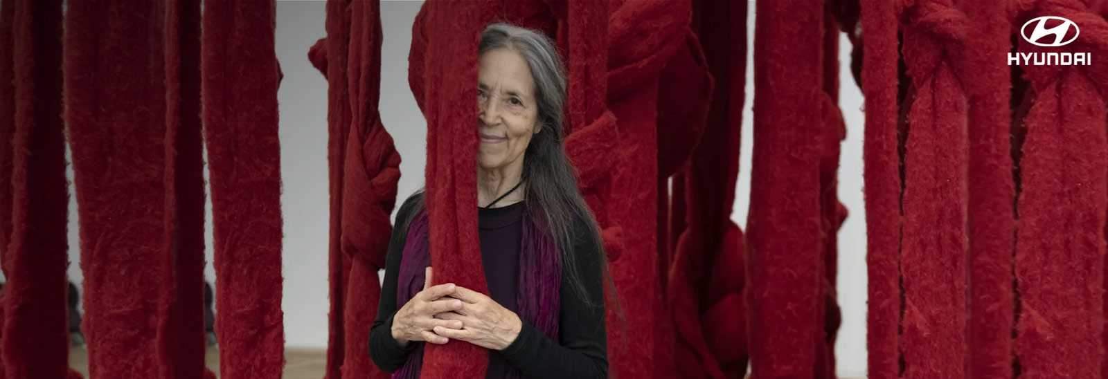 Cecilia Vicuña abrazando tiras de tela colgadas color rojo