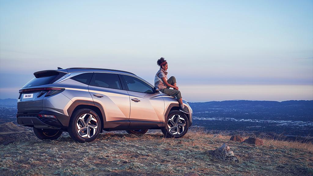 Mujer viendo hacia el horizonte sentada en cofre de Hyundai Tucson Híbrida color plata